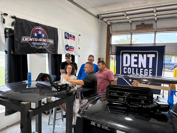A Dent College instructor leading a small hands-on paintless dent repair class, demonstrating dent removal on a vehicle panel while four students watch closely